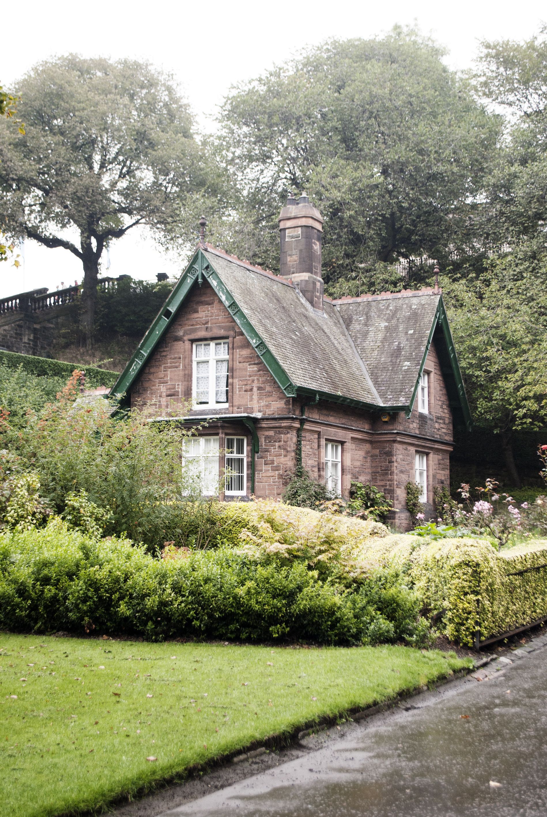 Kleines Natursteinhaus hinter einer geschnittenen Hecke, davor eine Rasenfläche
